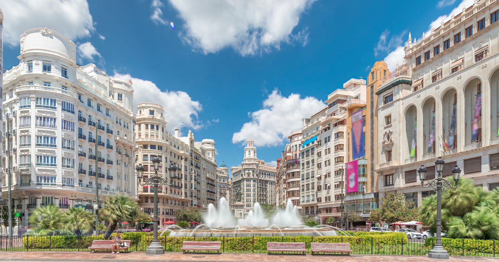 Panoramic view of a grand city square with a large central fountain, surrounded by ornate historic buildings, palm trees, and a clear blue sky with clouds overhead.