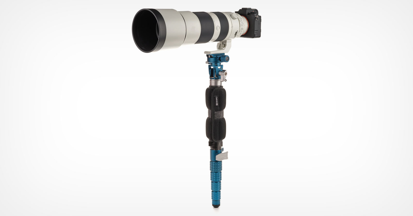 A professional camera with a large telephoto lens is mounted on a blue monopod. The camera is positioned horizontally, supported by the monopod, which features black and blue components and a padded grip. The background is plain white.