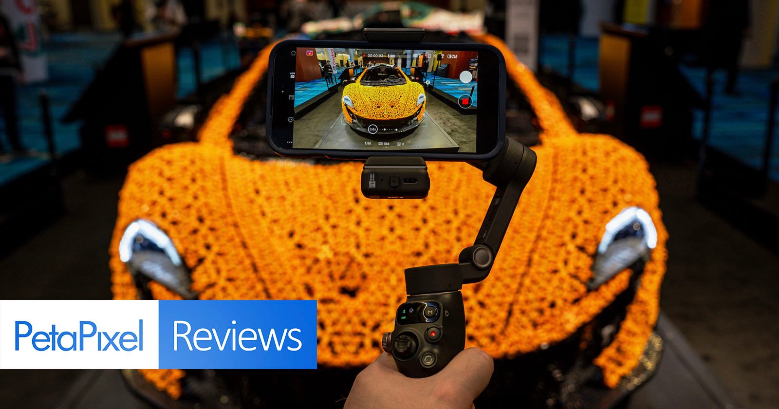 A person films a detailed LEGO car model using a smartphone mounted on a handheld gimbal. The LEGO car is bright orange and the setup suggests a tech or gadget review setting. PetaPixel Reviews is overlaid in the corner.