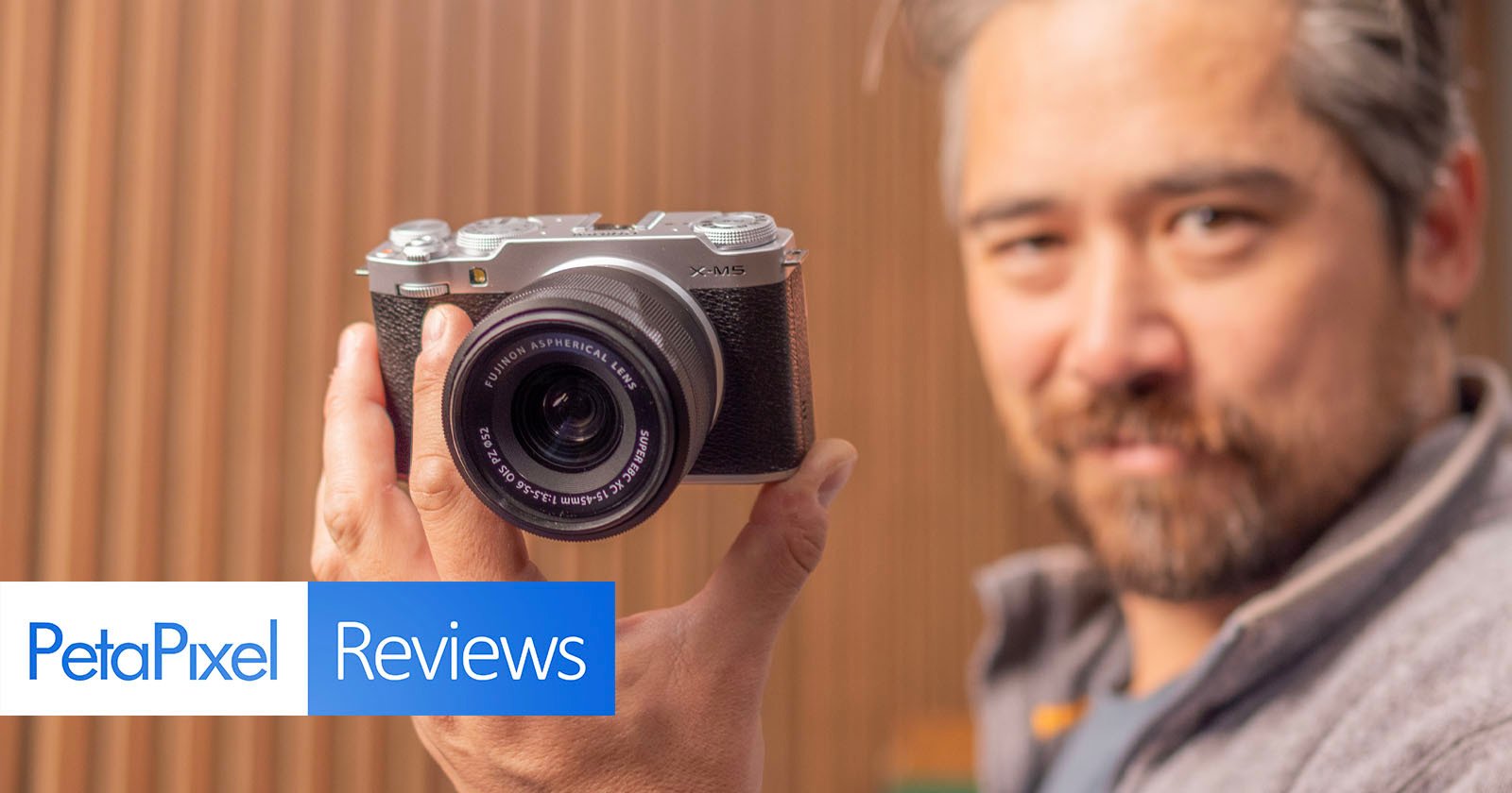 A person holding a vintage-style camera close to the lens, standing against a blurred wooden background. The image features the text PetaPixel Reviews in the bottom left corner.