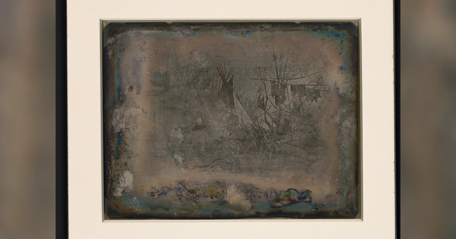 A faded, weathered daguerreotype photograph in a white frame shows faint images of trees and possibly a tent or hut, with darkened, tarnished edges giving the picture an aged appearance.