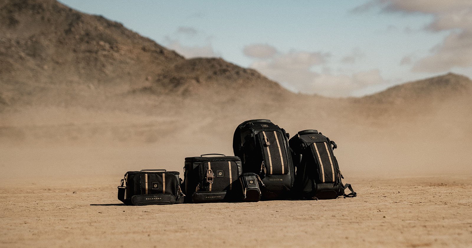 A lineup of various-sized black travel backpacks and bags is arranged in a desert landscape. Mountains and a clear blue sky are in the background, with a dusty ground in the foreground.