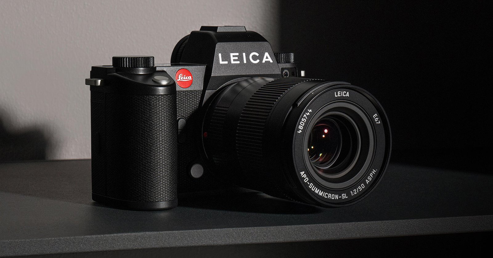 A black Leica camera with a 24-90mm zoom lens sits on a dark surface. The camera is partially illuminated from the left, emphasizing its textured grip and the iconic red Leica logo near the viewfinder.