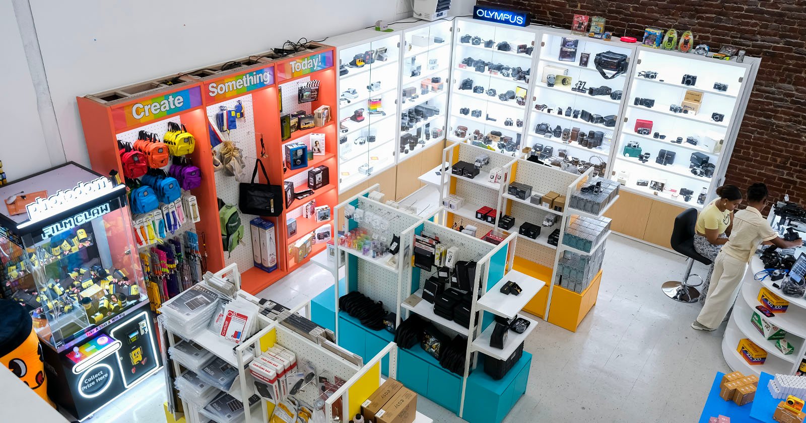 A brightly lit camera shop displays various cameras, lenses, and accessories on shelves. Two people sit at a counter on the right, while colorful bags and gear are organized on the left. The store has white floors and brick walls.