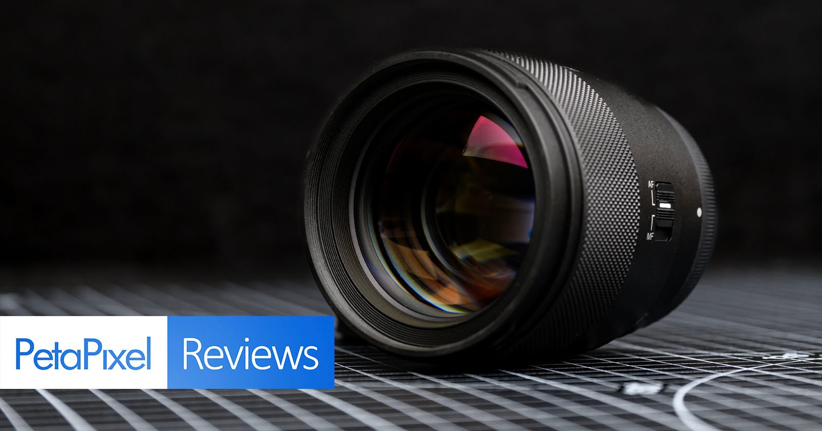 A camera lens is placed on its side on a grid-patterned surface, with PetaPixel Reviews text in the lower left corner against a dark, blurred background.
