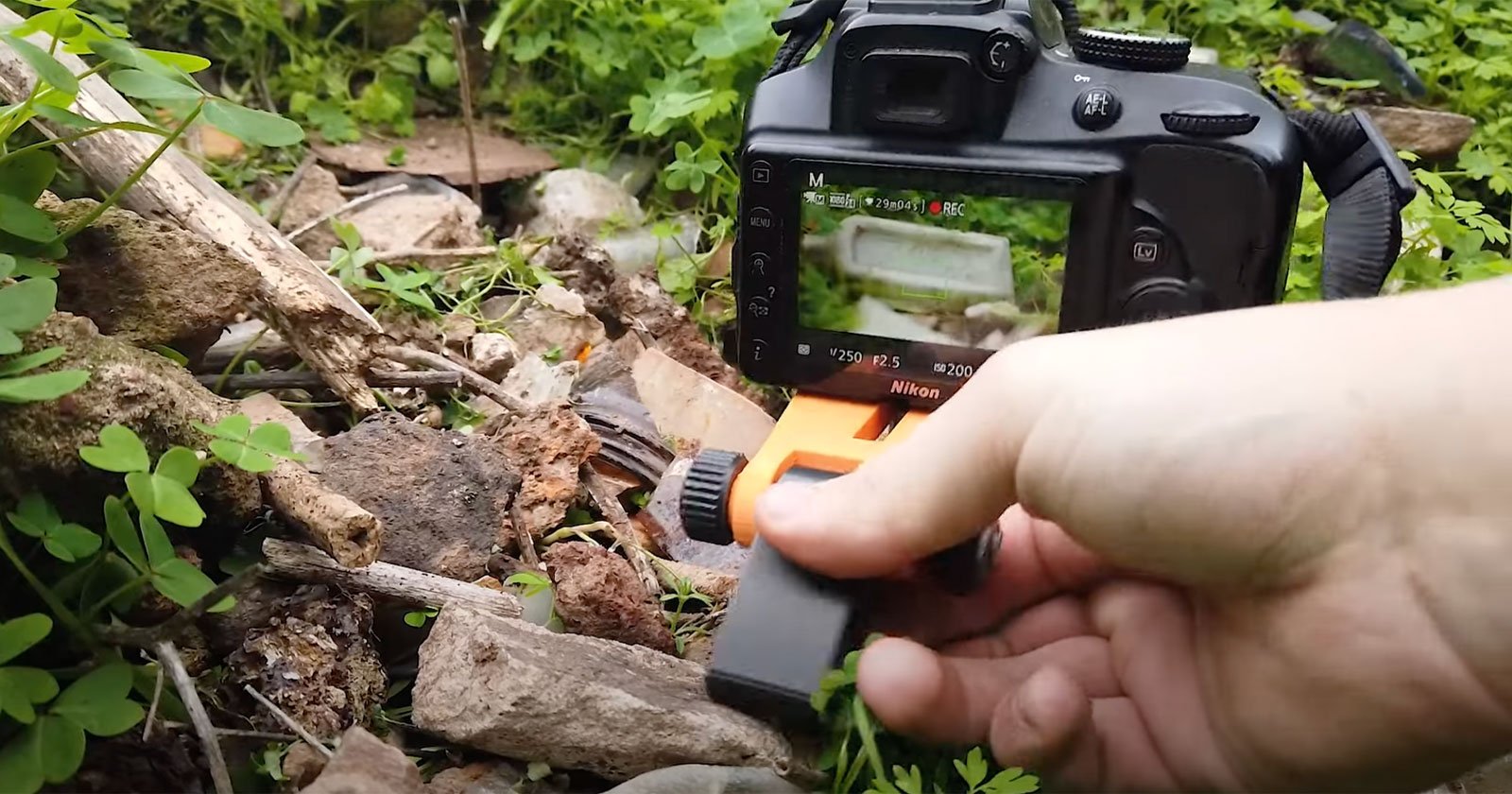 A person is using a Nikon DSLR camera to photograph an outdoor scene featuring rocks and greenery. The photographer's hand is adjusting a small orange and black tripod attached to the camera. The camera's screen displays the live view of the scene being captured.