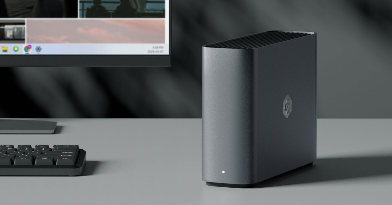A sleek, dark gray desktop computer tower sits on a desk next to a keyboard and a monitor, featuring a small white LED light and a logo on the front panel.