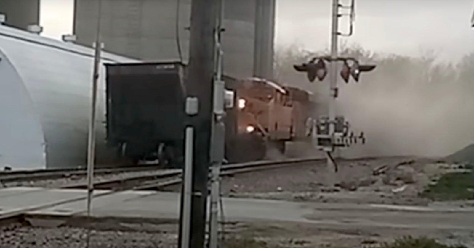 A freight train collides with a tractor-trailer at a railway crossing. Dust is seen billowing around the train and vehicle. A crossing signal is visible in the foreground, and an industrial building is on the left.