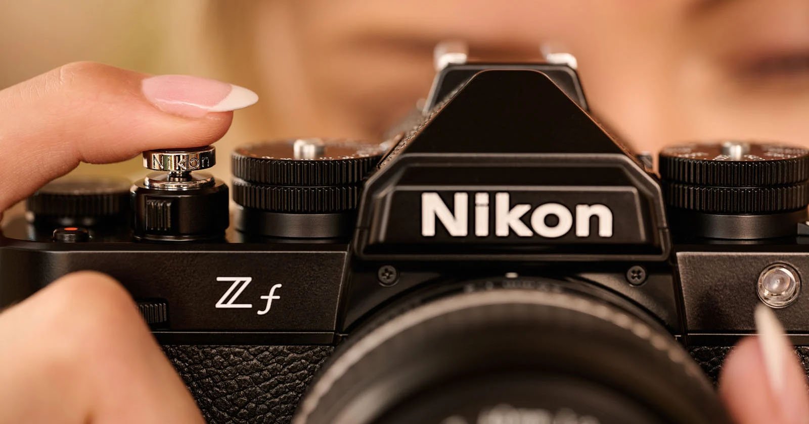 Close-up of a person holding a Nikon Zf camera, focusing on the camera's top controls and branding. The person's face is slightly blurred in the background, with their fingers adjusting the dial.