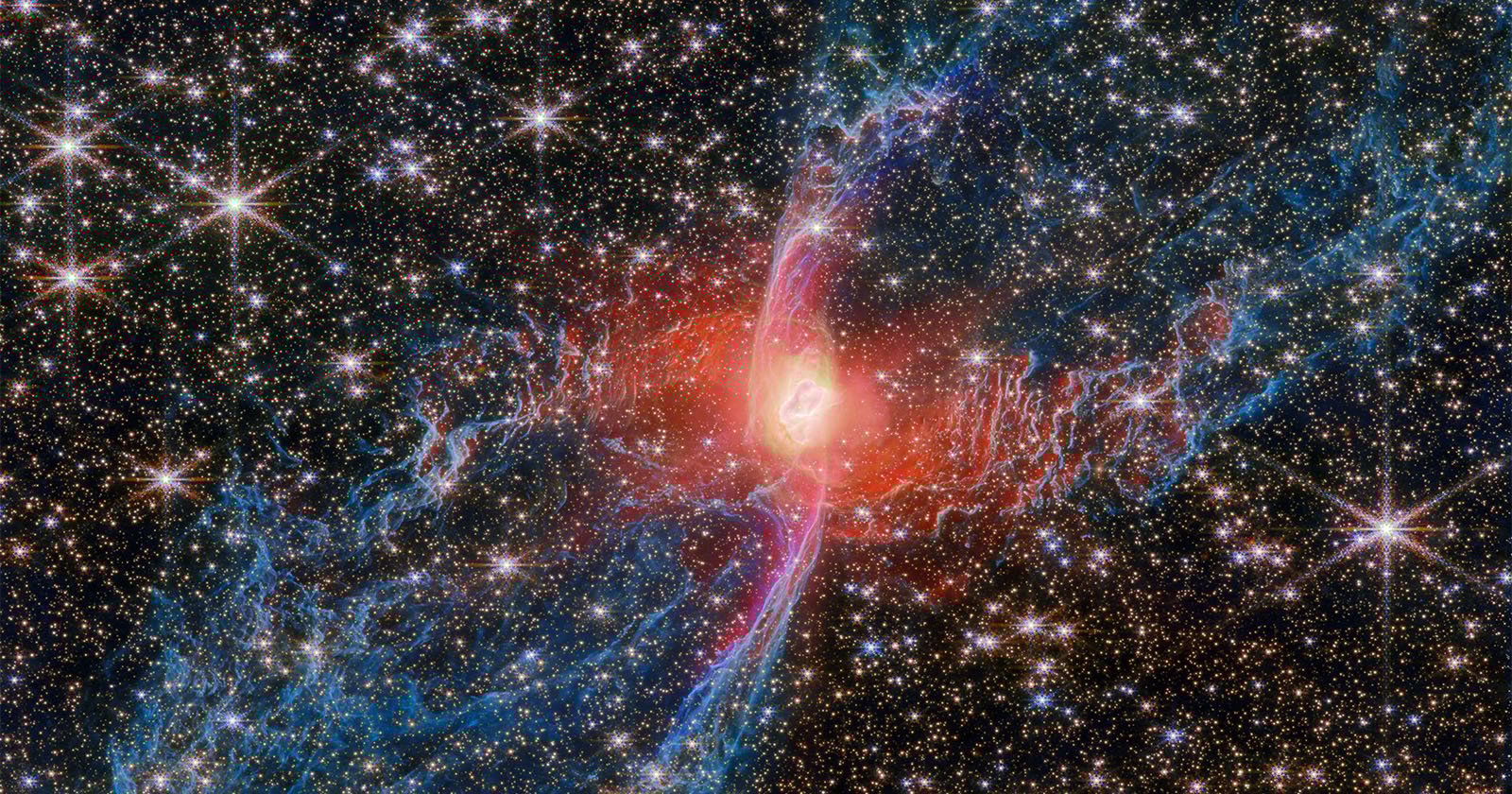 A vibrant nebula with a glowing red center and intricate blue and pink filaments stretches across a star-filled background in outer space. Many bright stars are scattered throughout the scene.