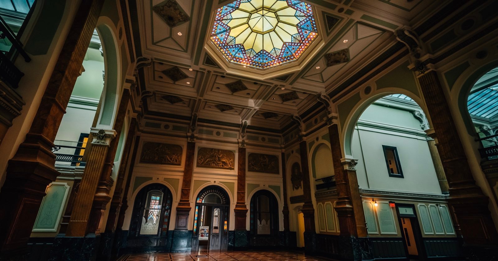 A grand hall with high ceilings, ornate columns, arched doorways, and a colorful stained glass skylight, illuminated by natural light streaming in. The architecture is classical and elegant.