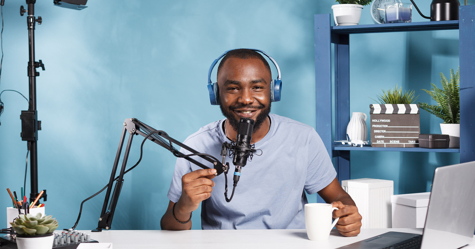 A man is sitting at a desk with a microphone and headphones, holding a white mug and smiling. The background is blue with a shelving unit holding plants, a clapperboard, and decorative items. Various electronic equipment is on the desk.