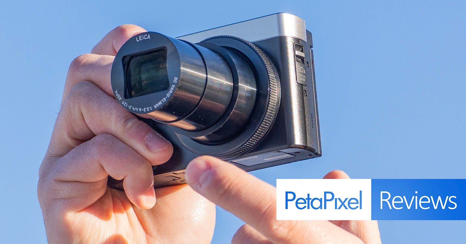 A person holding a compact camera with a zoom lens extended. The camera lens has Leica branding. The image has a blue sky background and features a PetaPixel Reviews graphic in the bottom right corner.