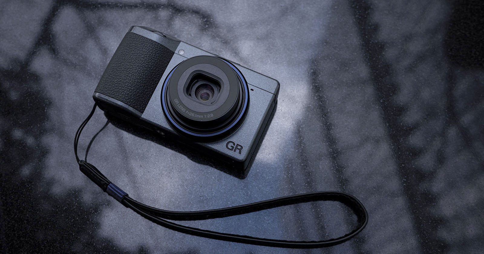 A compact digital camera with a black and gray design rests on a reflective surface. It features a visible lens and a wrist strap attached to the side. The surface has a blurred, abstract pattern in the background.