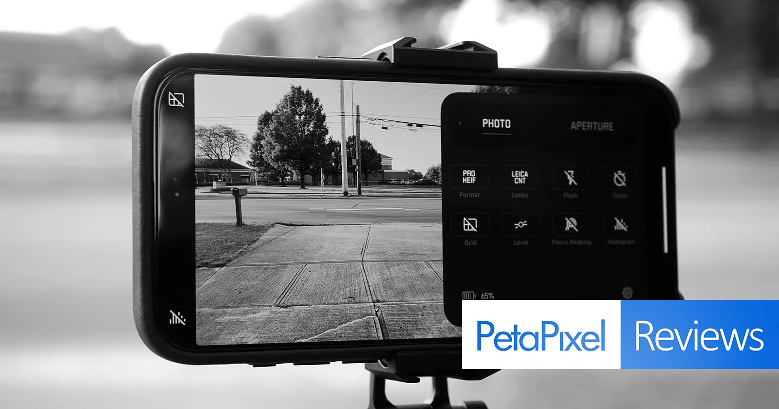A smartphone mounted on a tripod is taking a black-and-white photo of a street scene. Camera settings are visible on the screen. The PetaPixel Reviews logo is overlaid on the bottom right corner.