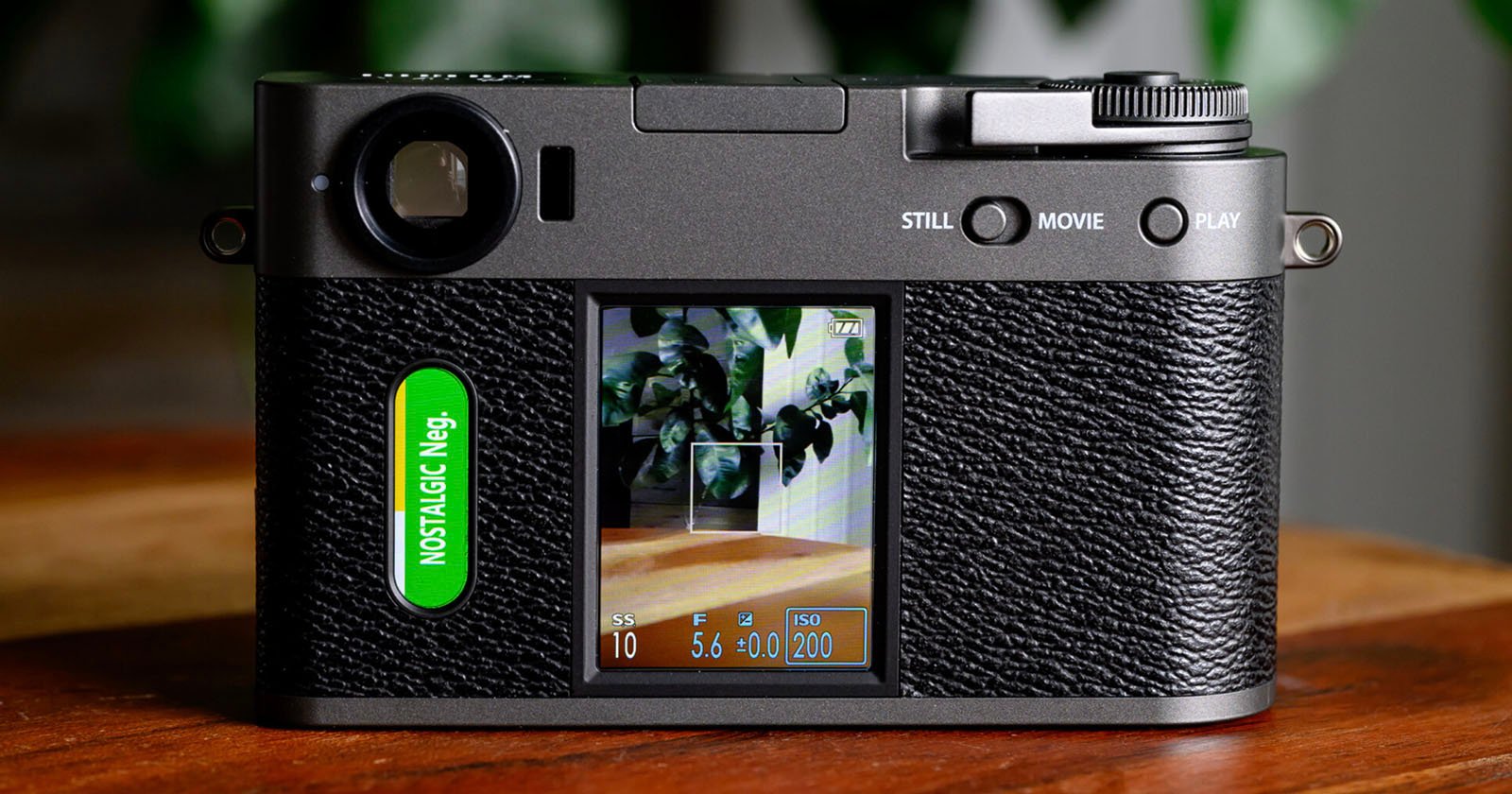 A close-up of the back of a digital camera with a textured black grip, displaying a screen showing a photo of leafy plants and camera settings, and a green button labeled NOSTALGIC Neg. on the left side.