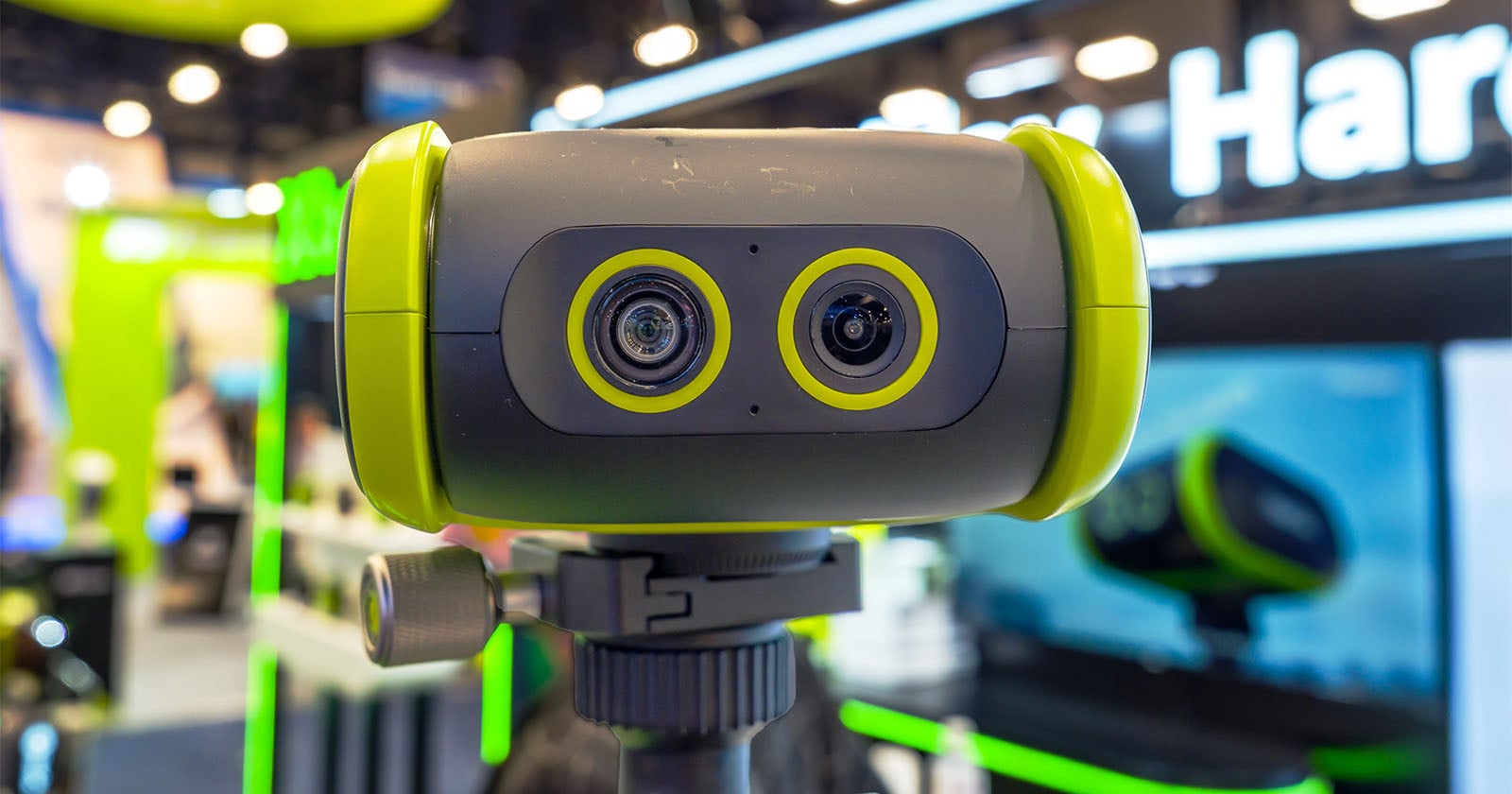 A close-up of a modern, dual-lens camera with green and black accents mounted on a tripod, displayed at a technology exhibition with blurred booths and signs in the background.