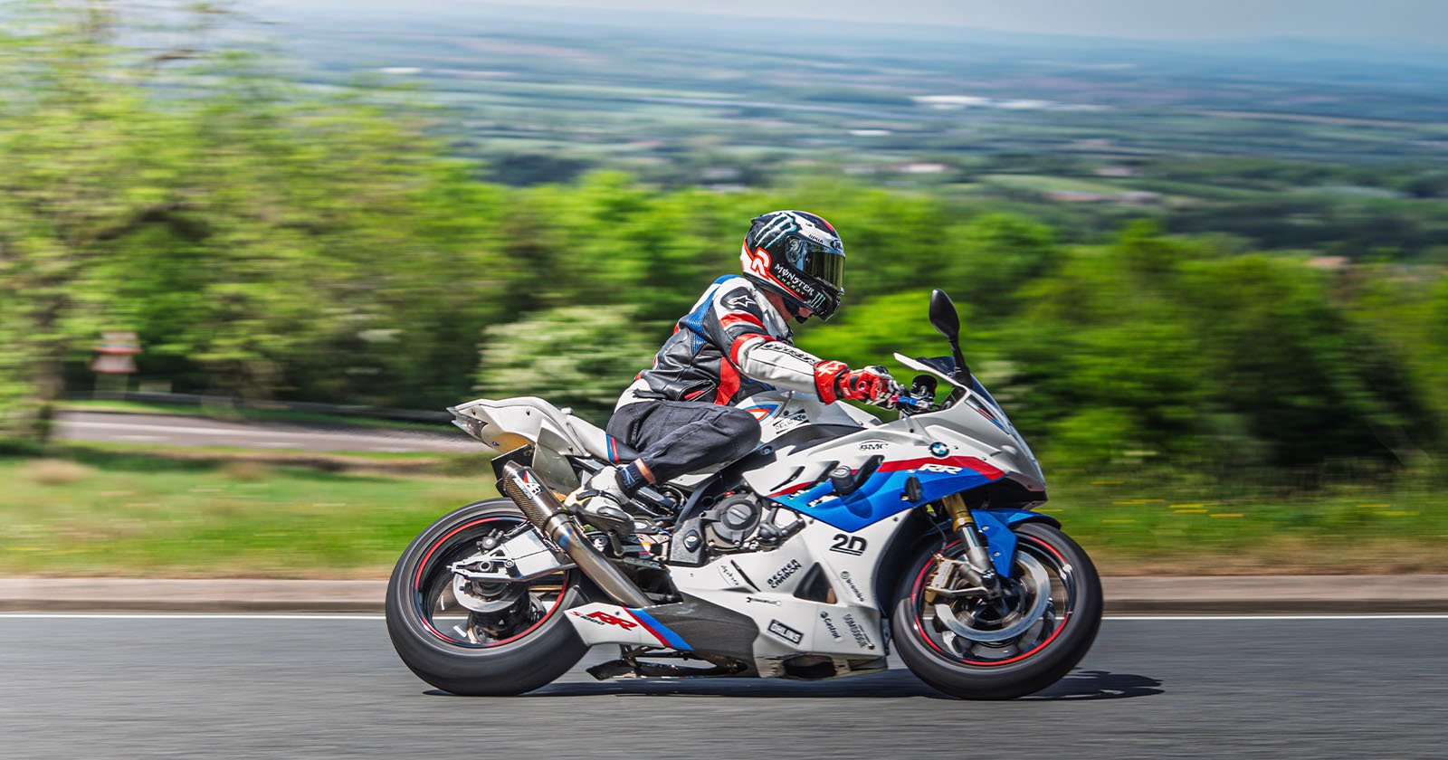 A motorcyclist in full gear rides a white sport bike with blue and red accents on a road, with a blurred green landscape in the background, suggesting high speed.