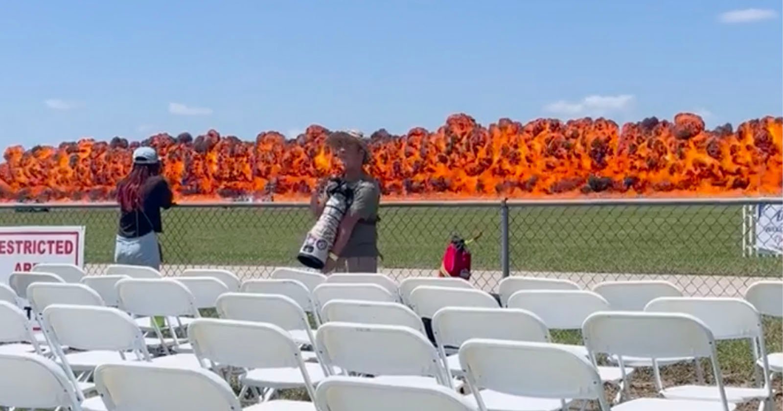 A row of empty white chairs faces a chain-link fence, behind which a massive explosion with bright orange flames occurs. Two people stand near the fence, one holding a large camera lens, both watching the blast.