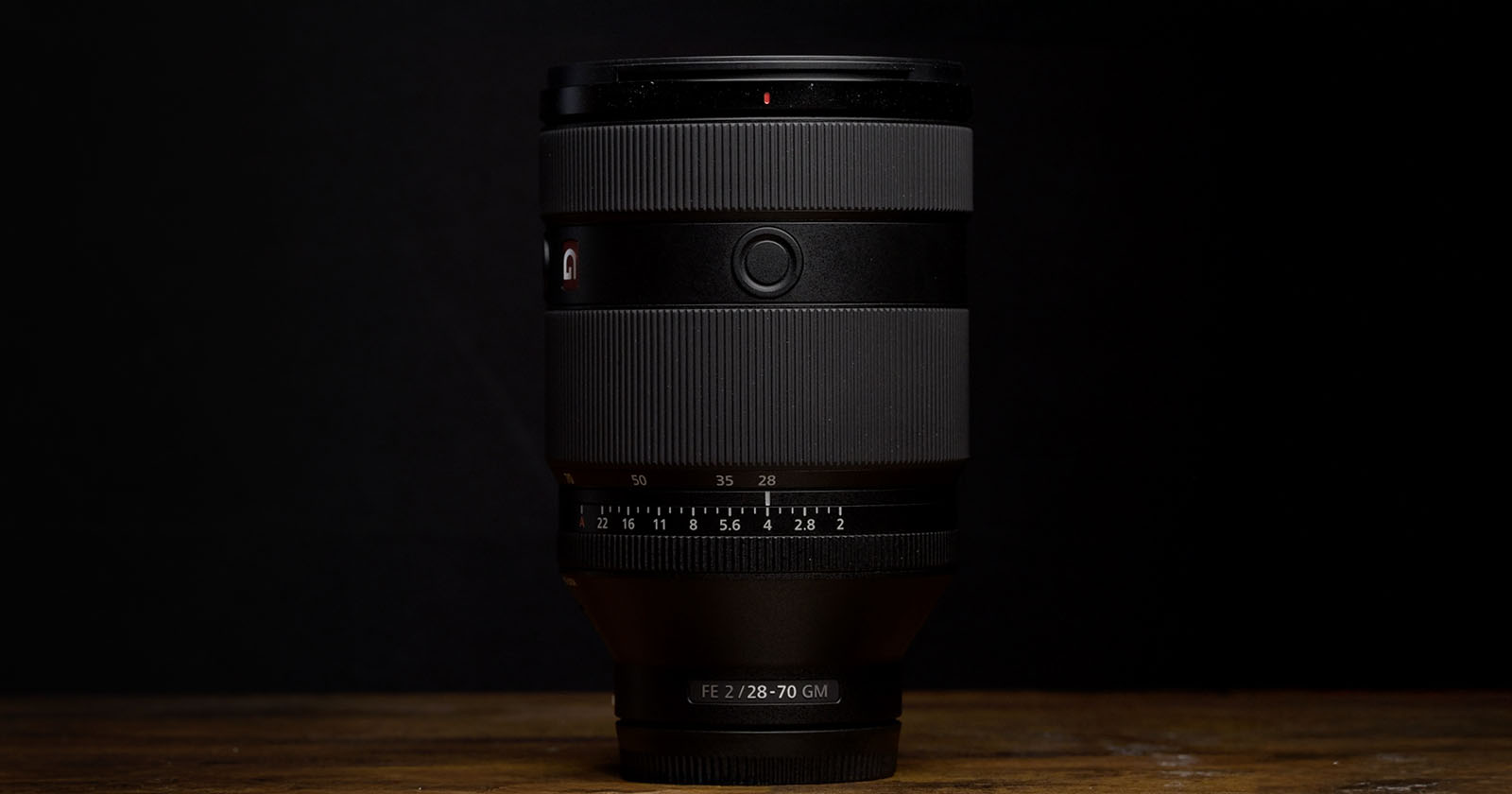 A camera lens stands upright on a wooden surface against a dark background. The lens features prominent zoom and focus rings with markings and a small red G logo.