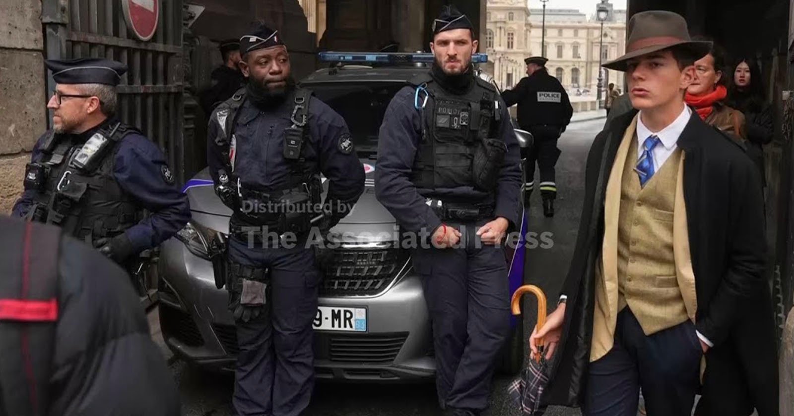 A group of police officers in uniform stand in front of a police car, while a man in a brown hat and suit with a yellow vest and umbrella walks past them. Several other people are in the background.