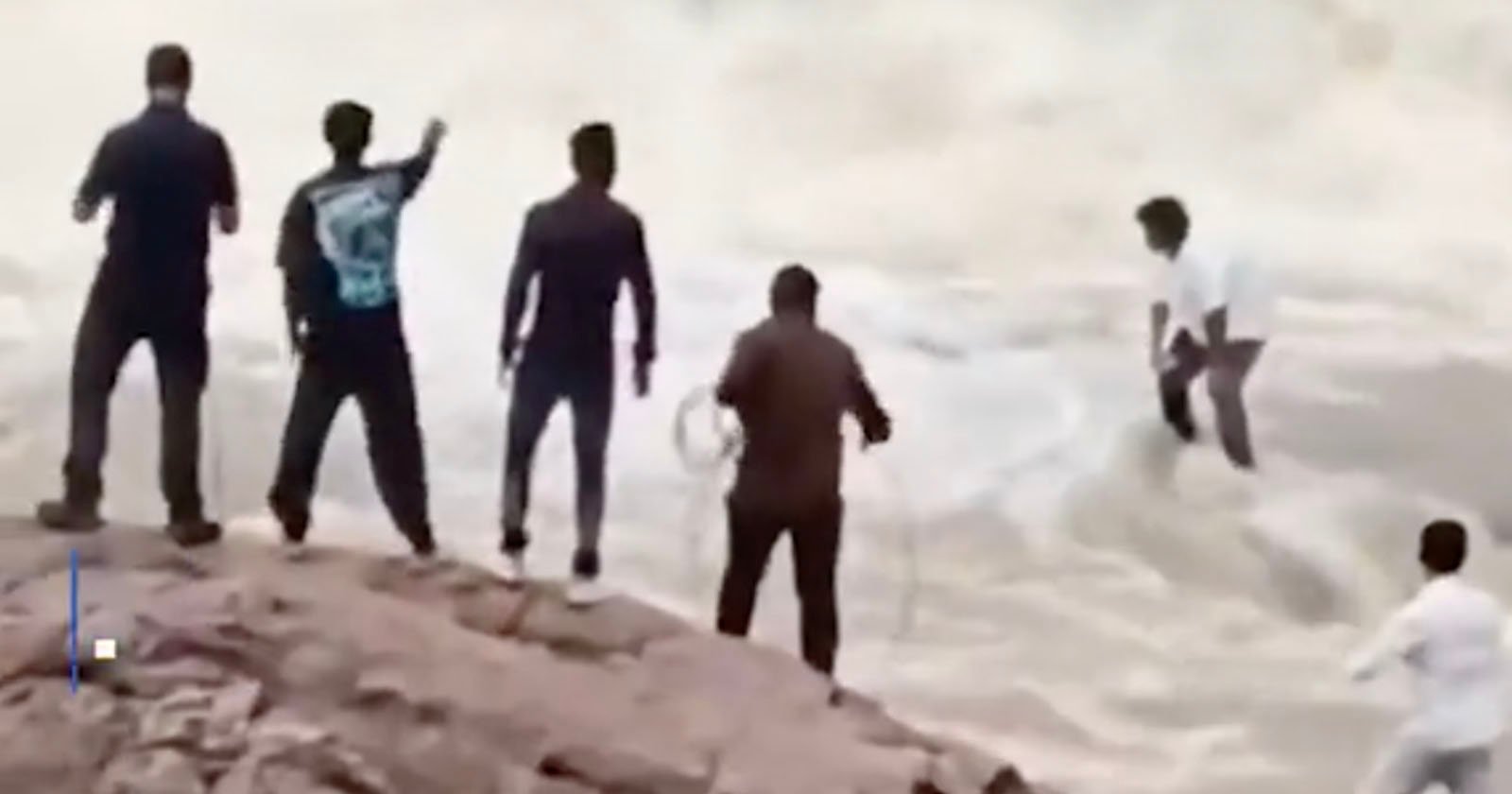 Five men stand on a rocky riverbank, some holding ropes, while one man is in the water. The scene appears tense, suggesting a possible rescue attempt in progress. The water is rough and fast-moving.