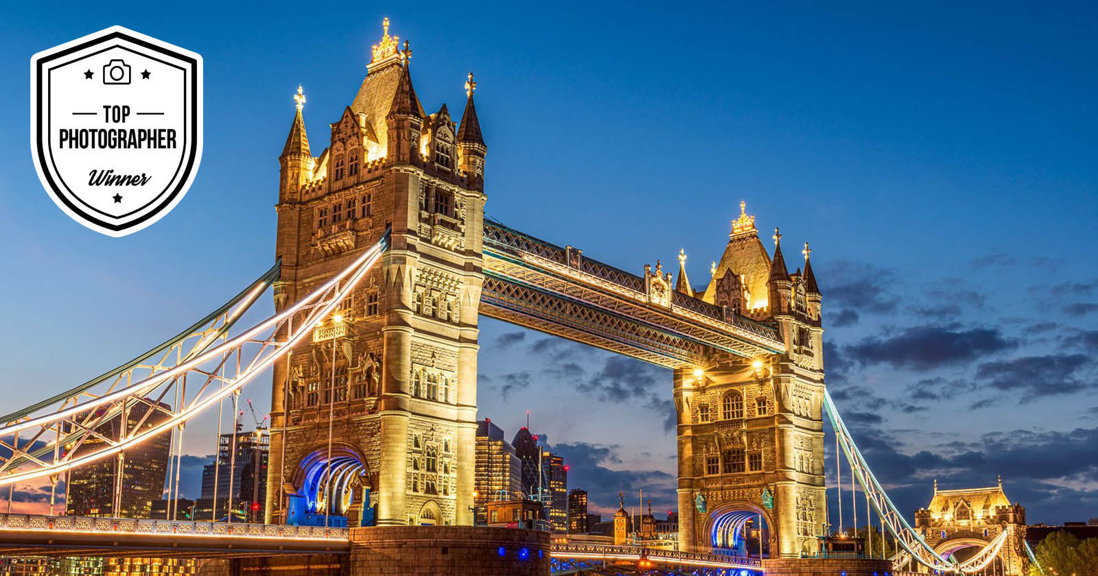 A beautifully illuminated Tower Bridge at dusk, with a Top Photographer Winner badge displayed in the top left corner. The bridge's lights shine against a backdrop of a darkening sky and city skyline.