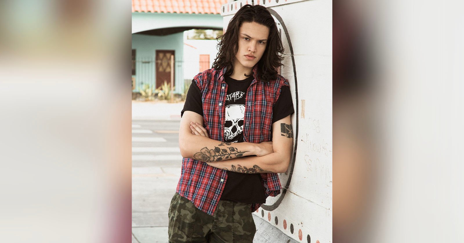 Young person with long dark hair, wearing a red plaid sleeveless shirt over a black graphic t-shirt and camouflage pants, stands with arms crossed against a white wall outside. Visible tattoos on arms.