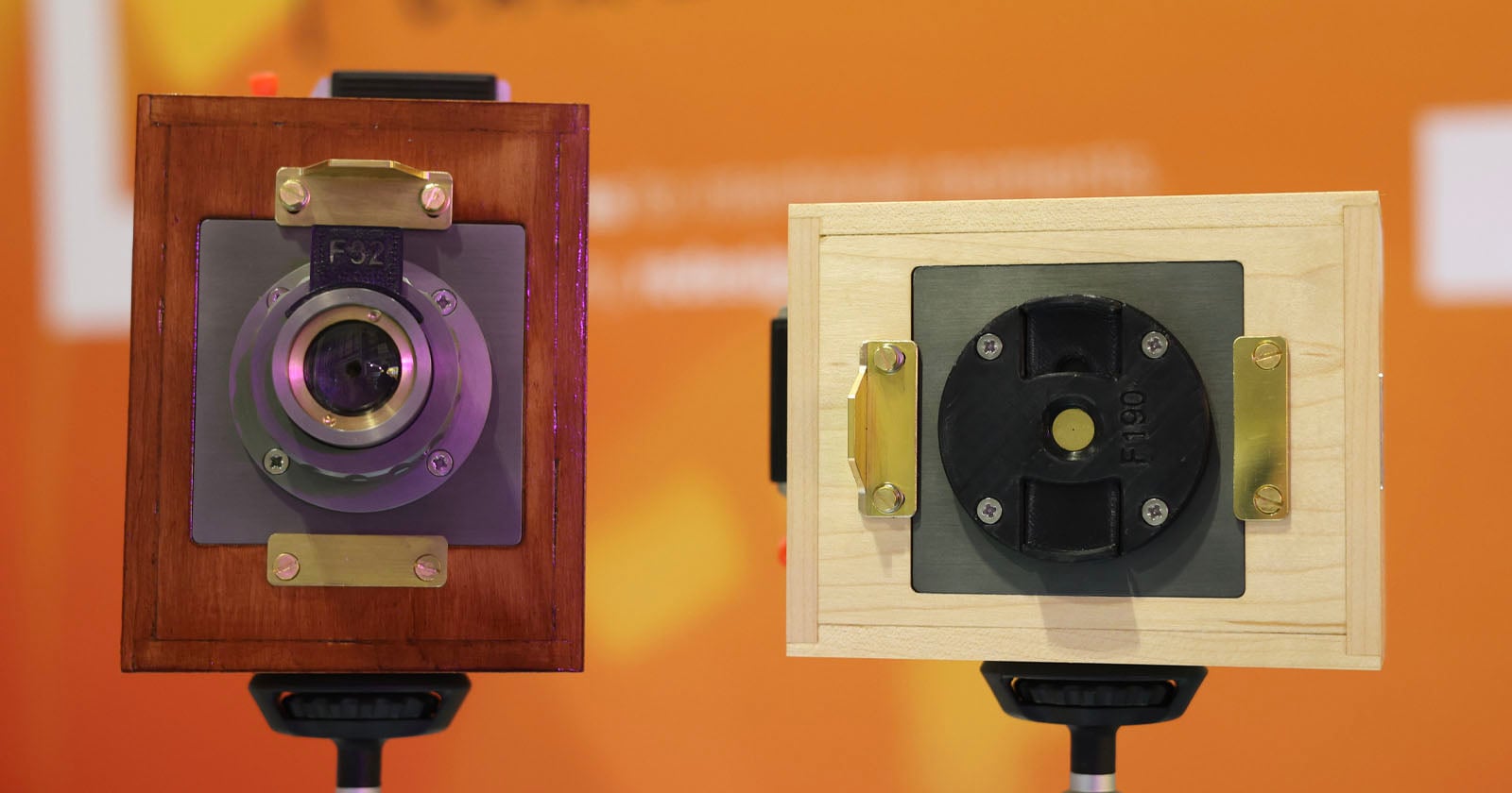 Two vintage-style box cameras with wooden bodies—one in dark wood, one in light wood—are mounted on tripods against an orange, blurred background.