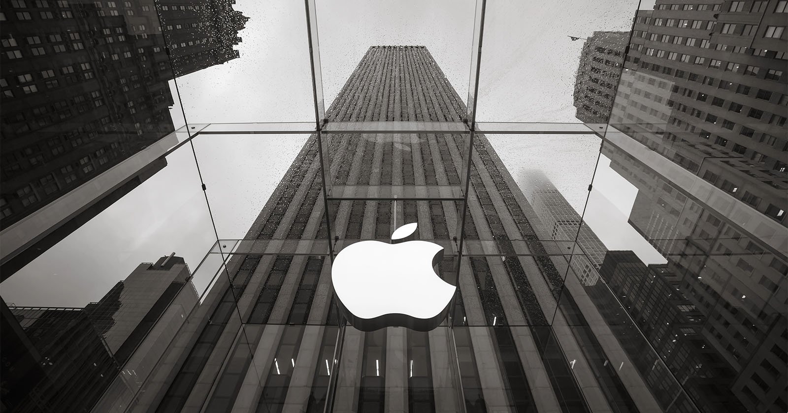 A monochrome photo of the Apple Store entrance featuring a large illuminated Apple logo on glass, with tall city buildings and a cloudy sky reflected in the background.