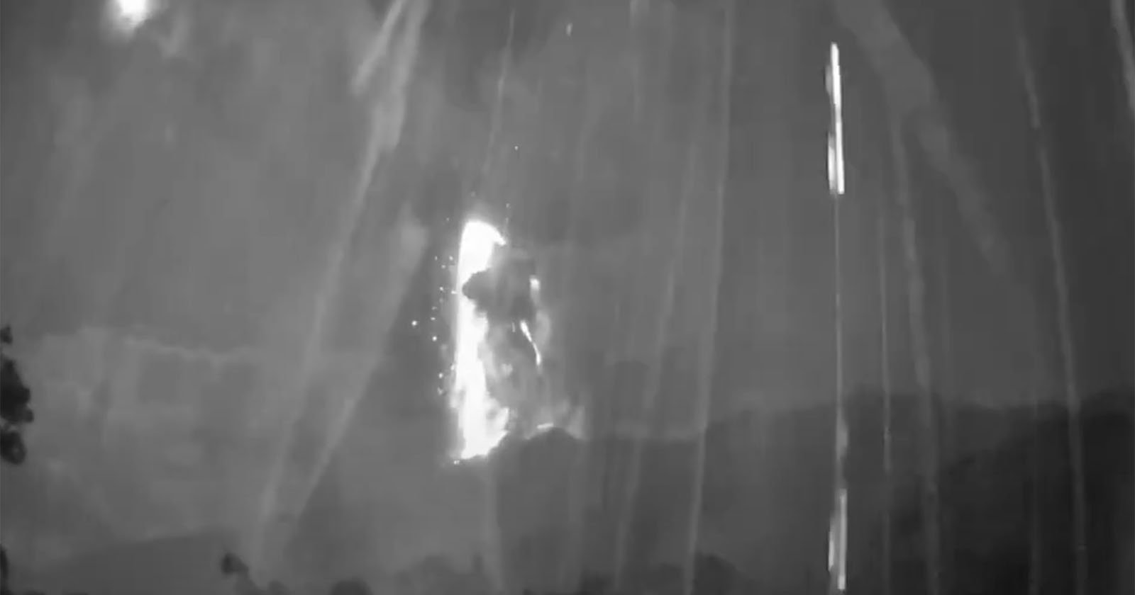 A black and white image shows a bright eruption or explosion with sparks and smoke against a dark sky, partially obscured by streaks of light, possibly from rain or camera artifacts. Trees and mountains are silhouetted below.