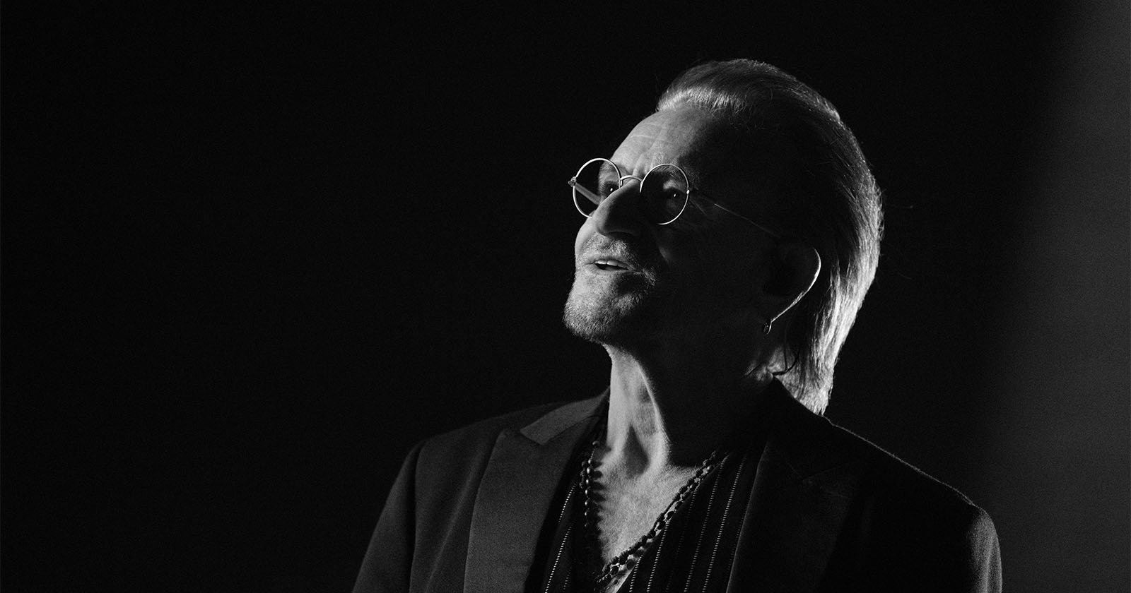 A man wearing glasses and a suit jacket, shown in black and white, looks upward with a contemplative expression. The background is dark, creating dramatic lighting on his face and hair.
