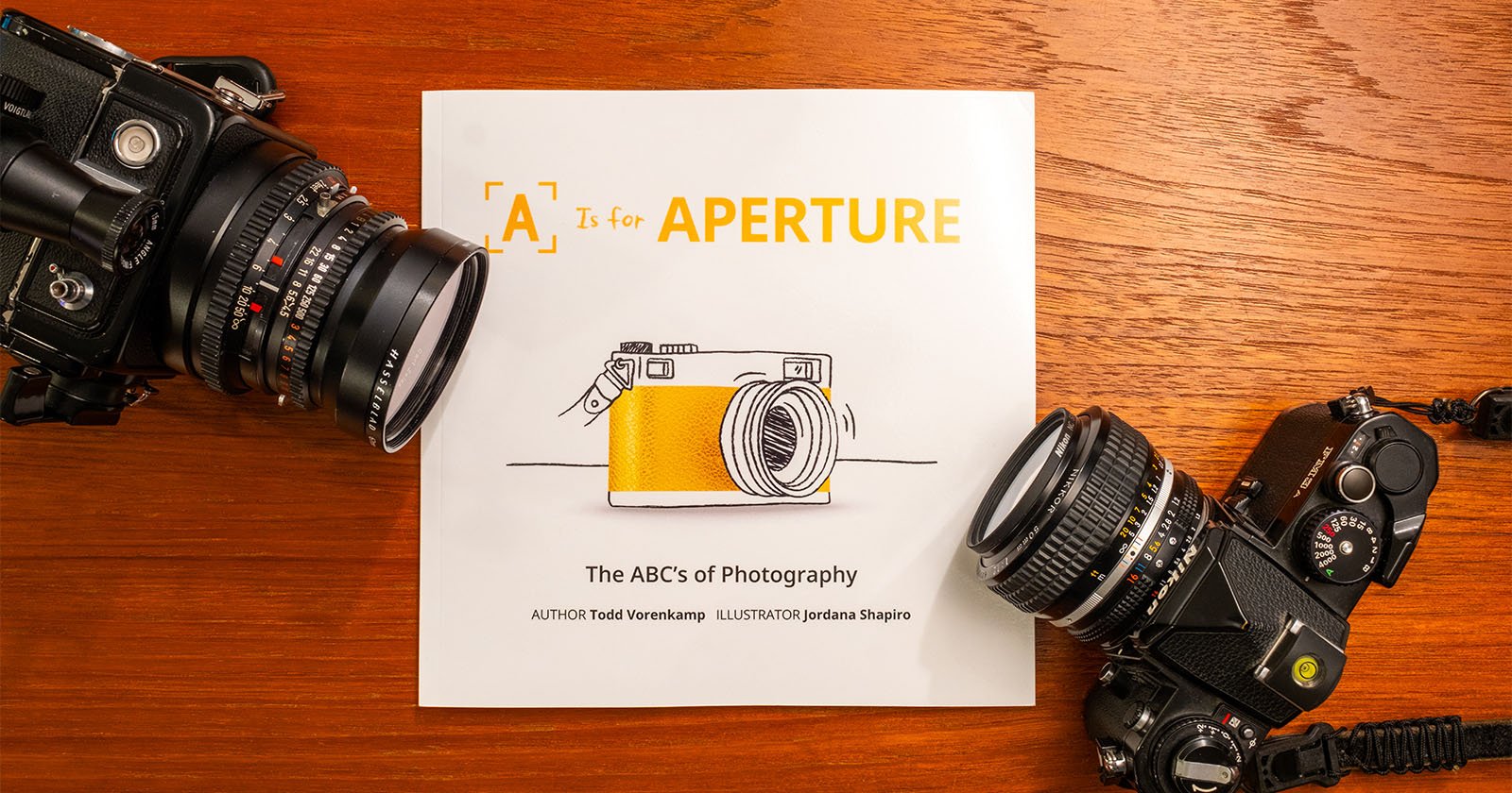 A book titled A is for Aperture: The ABC's of Photography by Todd Vorenkamp, illustrated by Jentana Shapiro, rests on a wooden surface. Two vintage cameras flank the book, one on the left and one on the right.