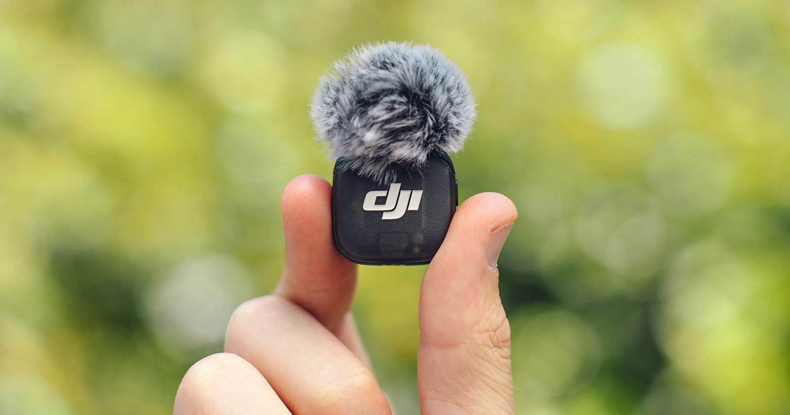 A hand holds a small DJI microphone with a gray furry windscreen against a blurred green background.