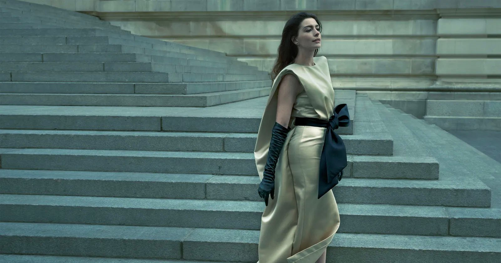 A woman in a cream satin dress with a large black bow and long black gloves stands near wide stone steps, gazing pensively to the side. The background features a grand, gray stone building.