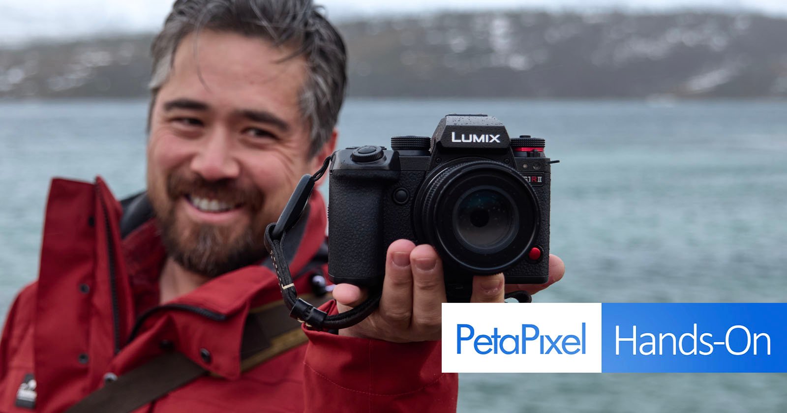 A person wearing a red jacket holds a Lumix camera towards the viewer, with a blurred body of water and snowy landscape in the background. The text PetaPixel Hands-On is displayed on the right side of the image.