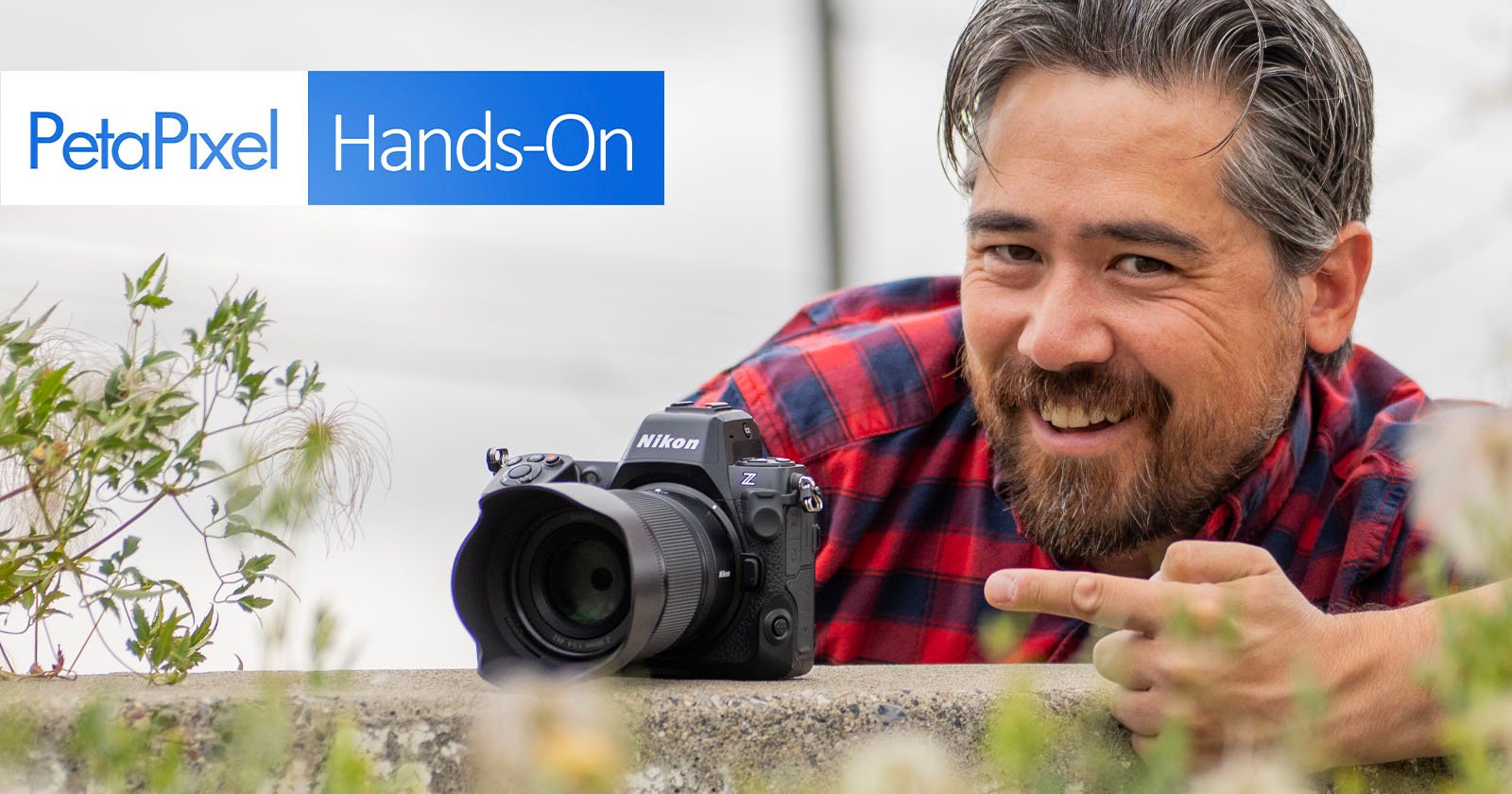 A person with a beard, wearing a red and black checkered shirt, leans over a wall and points to a Nikon camera placed on it. The text PetaPixel Hands-On is visible in the top left corner of the image.