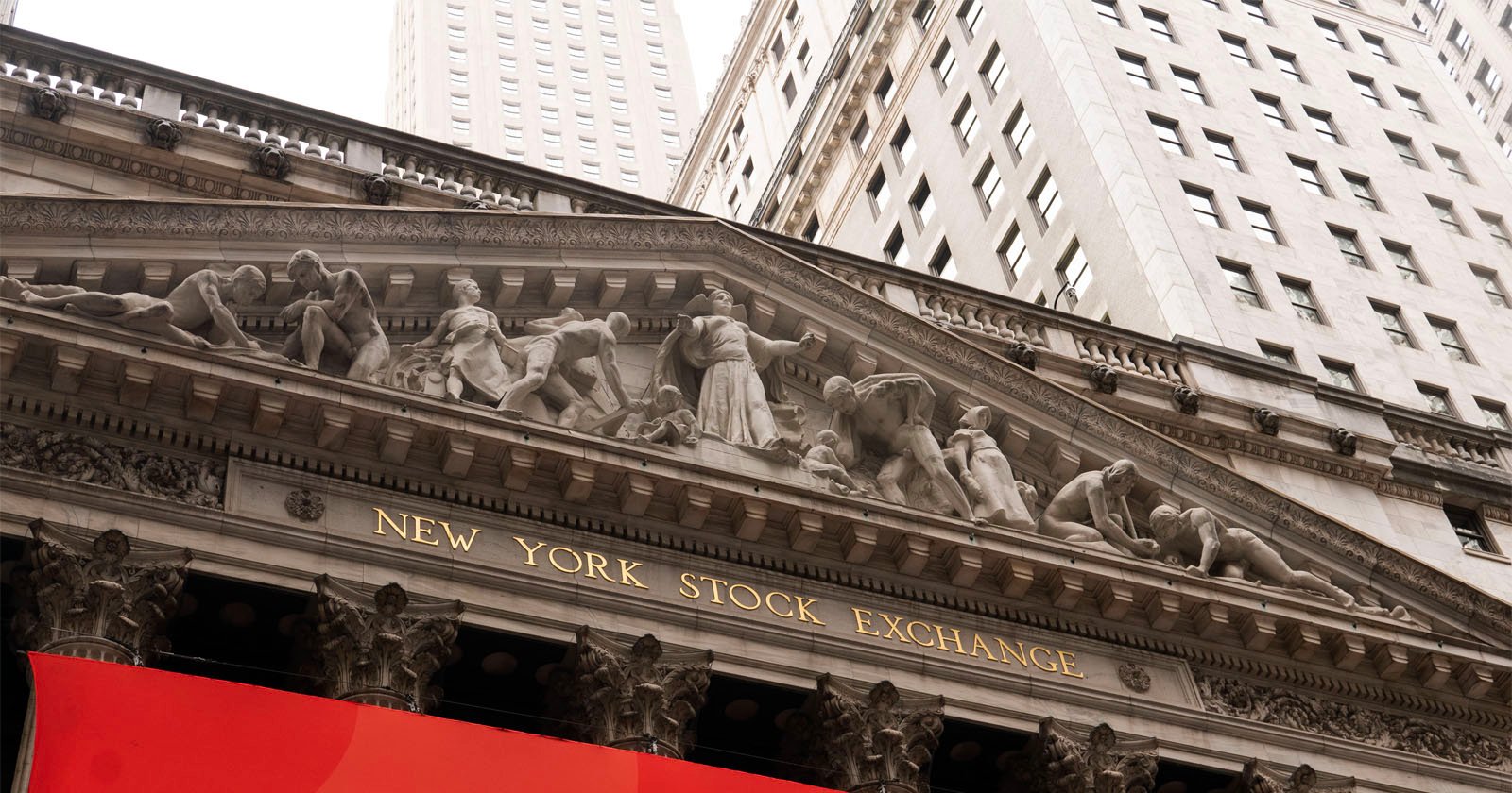 The facade of the New York Stock Exchange building, featuring classical columns and sculpted figures above the entrance, with tall skyscrapers in the background.