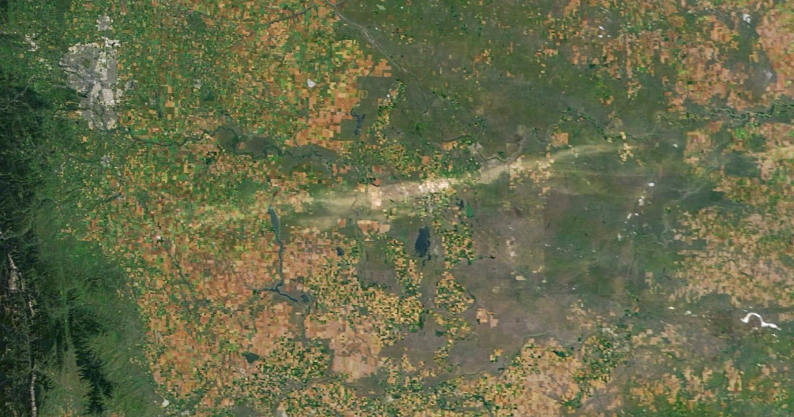 A satellite image shows a patchwork of farmland with various shades of green and brown, interspersed with small lakes and rivers. A pale diagonal stripe crosses the center. A city is visible in the upper left corner.