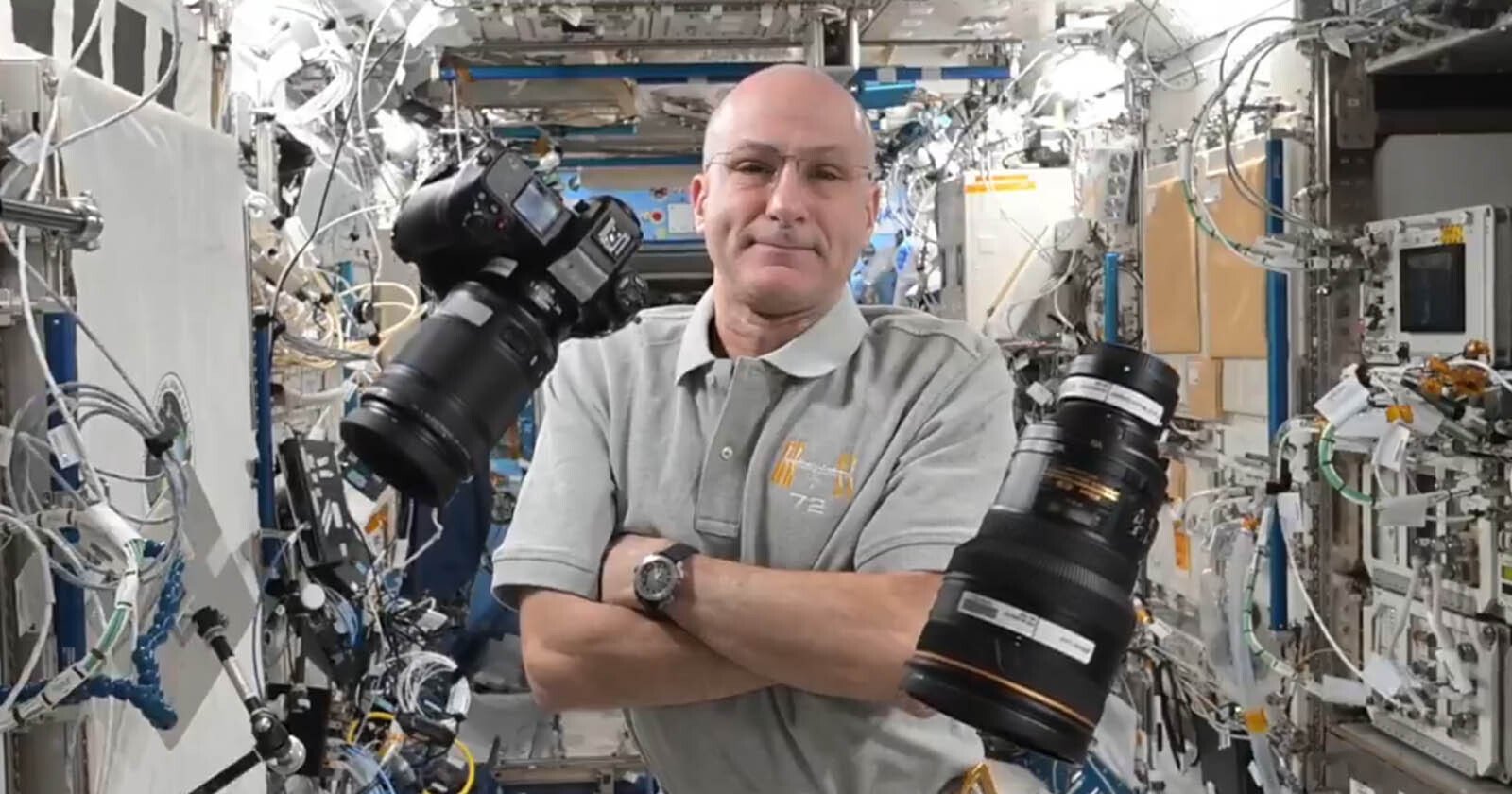 An astronaut in a gray shirt folds his arms inside a space station, with cameras floating nearby. He stands amid an array of equipment and cables, highlighting the zero-gravity environment.
