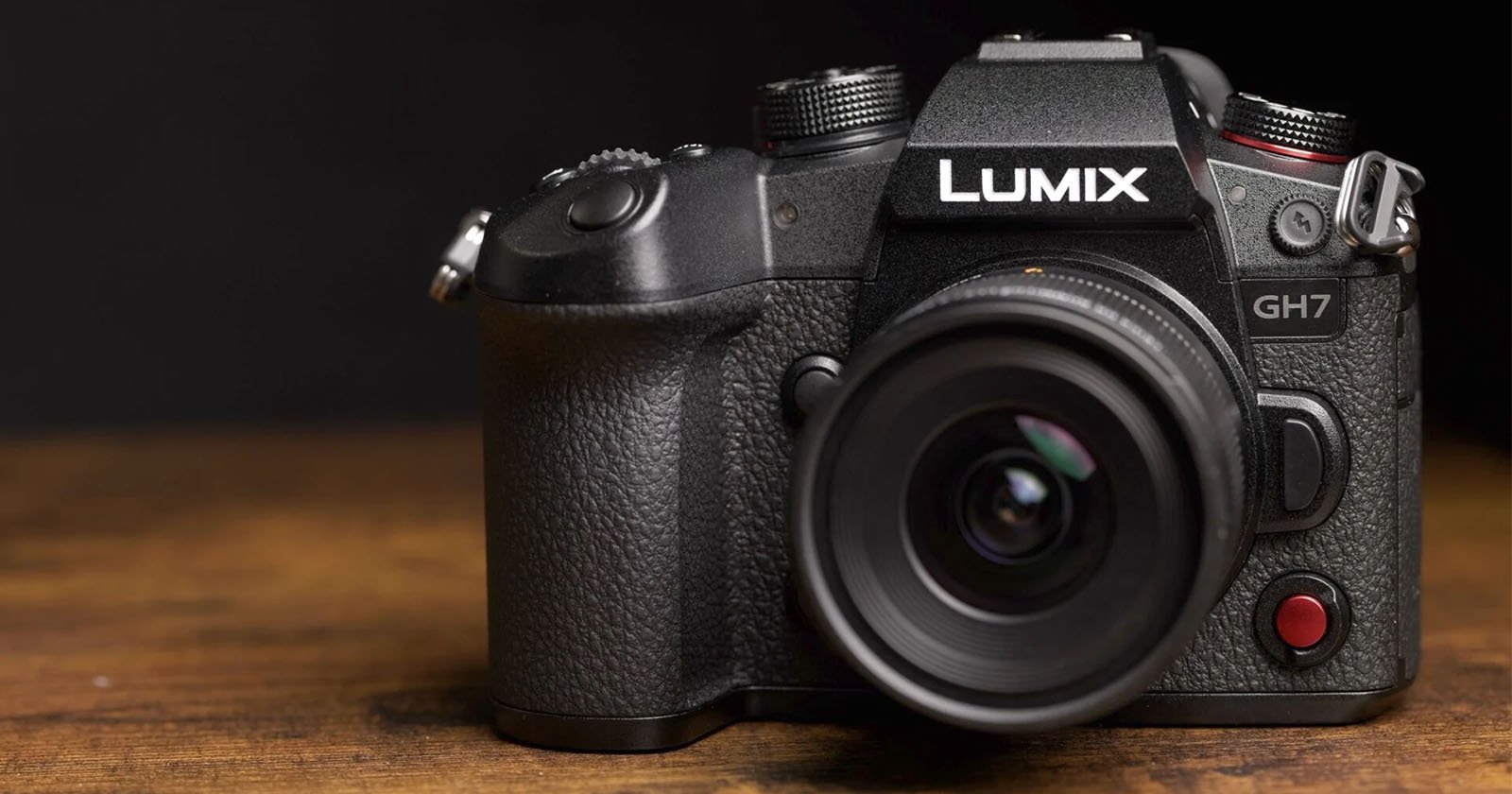 A black Panasonic LUMIX GH7 camera with a wide-angle lens is placed on a wooden surface, against a dark background. The camera has a textured grip, various buttons, and a large viewfinder. The brand name LUMIX is clearly visible above the lens.