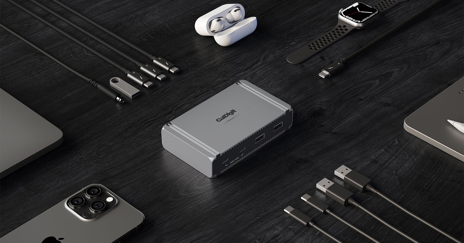 A variety of tech accessories arranged on a dark surface, including a smartwatch, earphones in a charging case, a smartphone, cables, a laptop, and a compact gray device at the center.