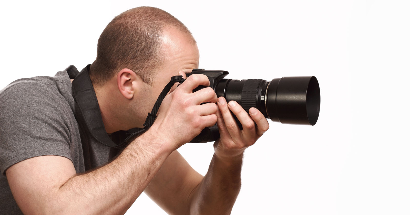 A person in a grey shirt is holding a DSLR camera with a long zoom lens up to their eye, capturing a photo. The person has short hair and uses a camera strap around their neck. The background is plain white.