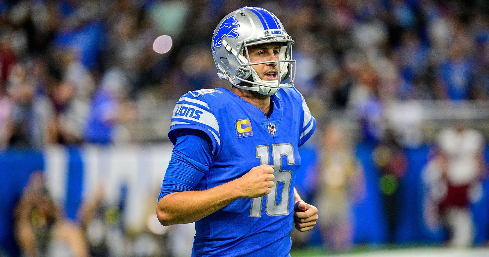 A Detroit Lions football player wearing a blue uniform and helmet with the number 16 on his jersey runs on the field during a game. The crowd is blurred in the background.