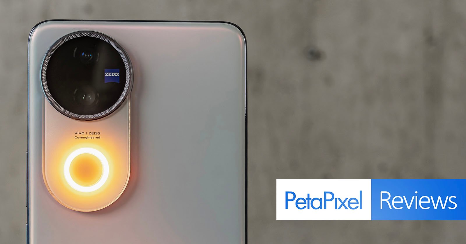 Close-up of a smartphone's rear camera with a prominent circular camera unit featuring a Zeiss lens. A ring light around the camera glows orange. The text PetaPixel Reviews is displayed in the bottom right corner on a gray background.