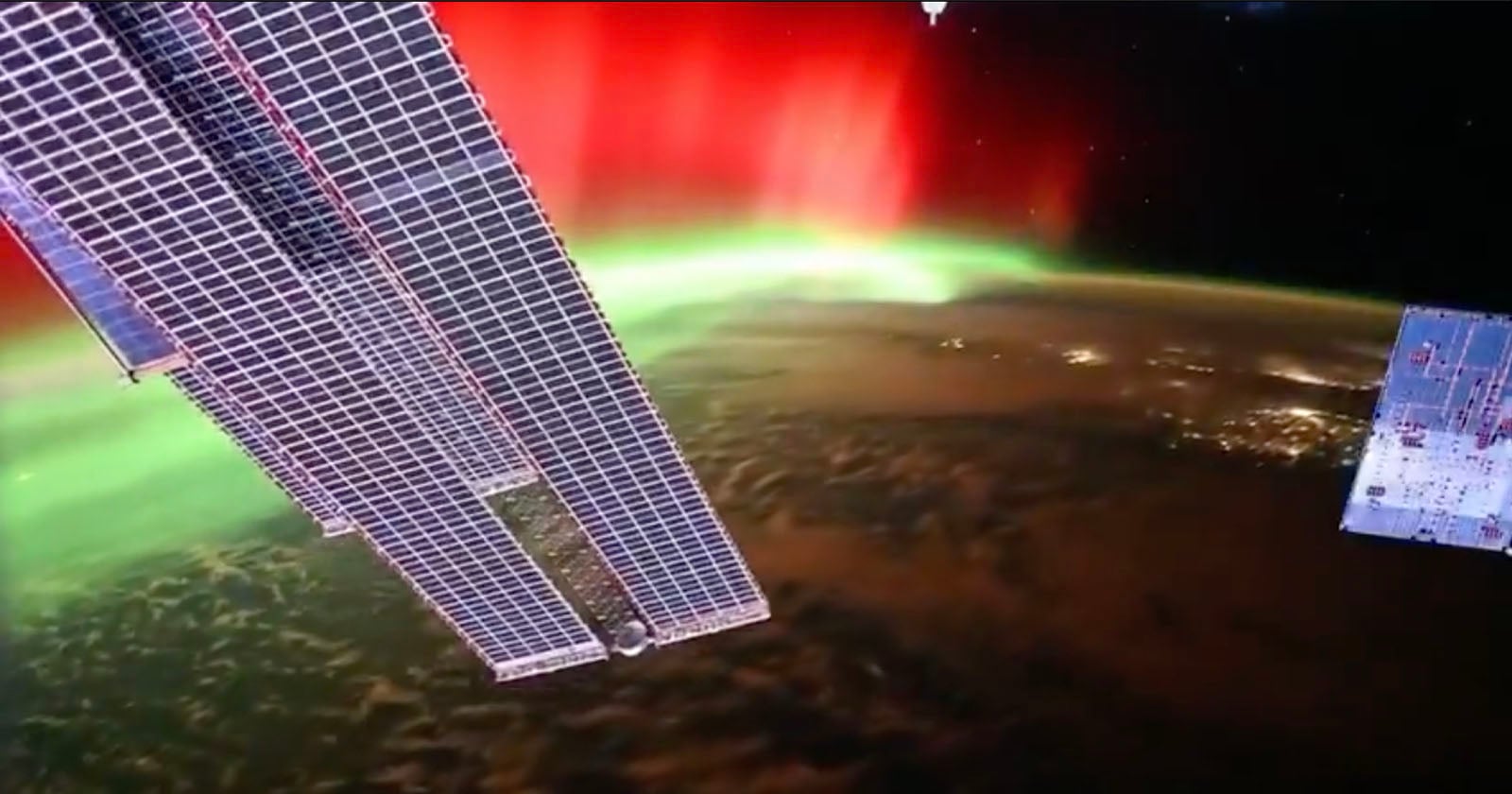 View from space showing the edge of a solar panel and part of the International Space Station with a vivid red and green aurora over the Earth’s surface, and city lights visible below.