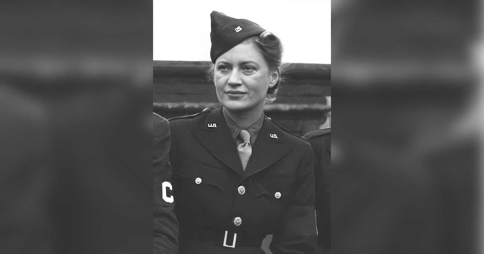Black and white photo of a woman in a military uniform, including a peaked cap. She has a confident expression and is pictured from the waist up. The uniform features insignia and buttons, and she is posed with other partially visible, similarly attired individuals.
