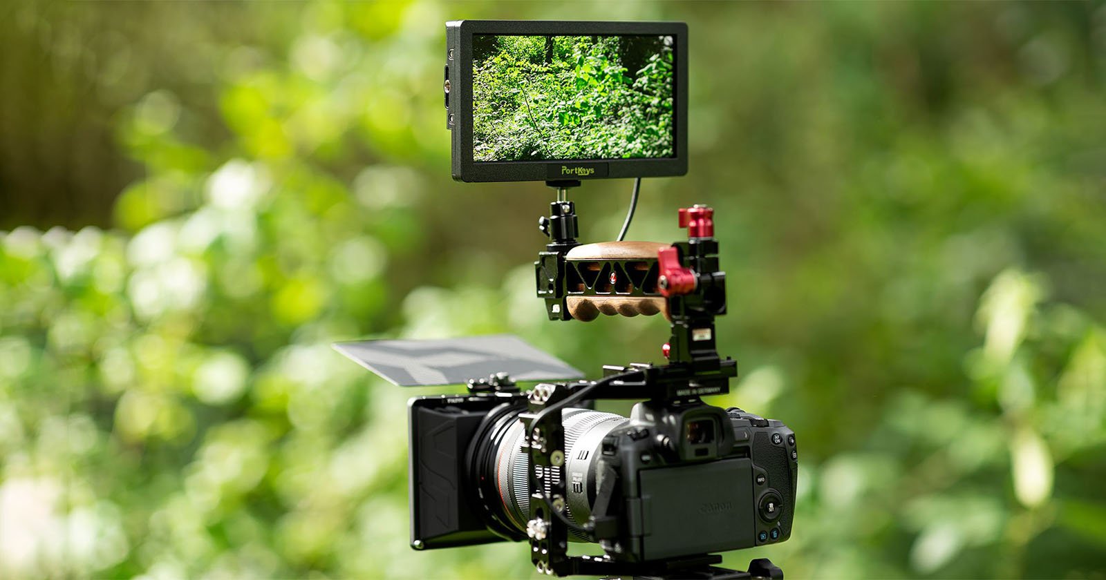 A DSLR camera mounted on a tripod in an outdoor setting. Attached to the camera is a field monitor displaying the camera's view of lush green foliage in the background. The setup appears to be for professional videography or photography.