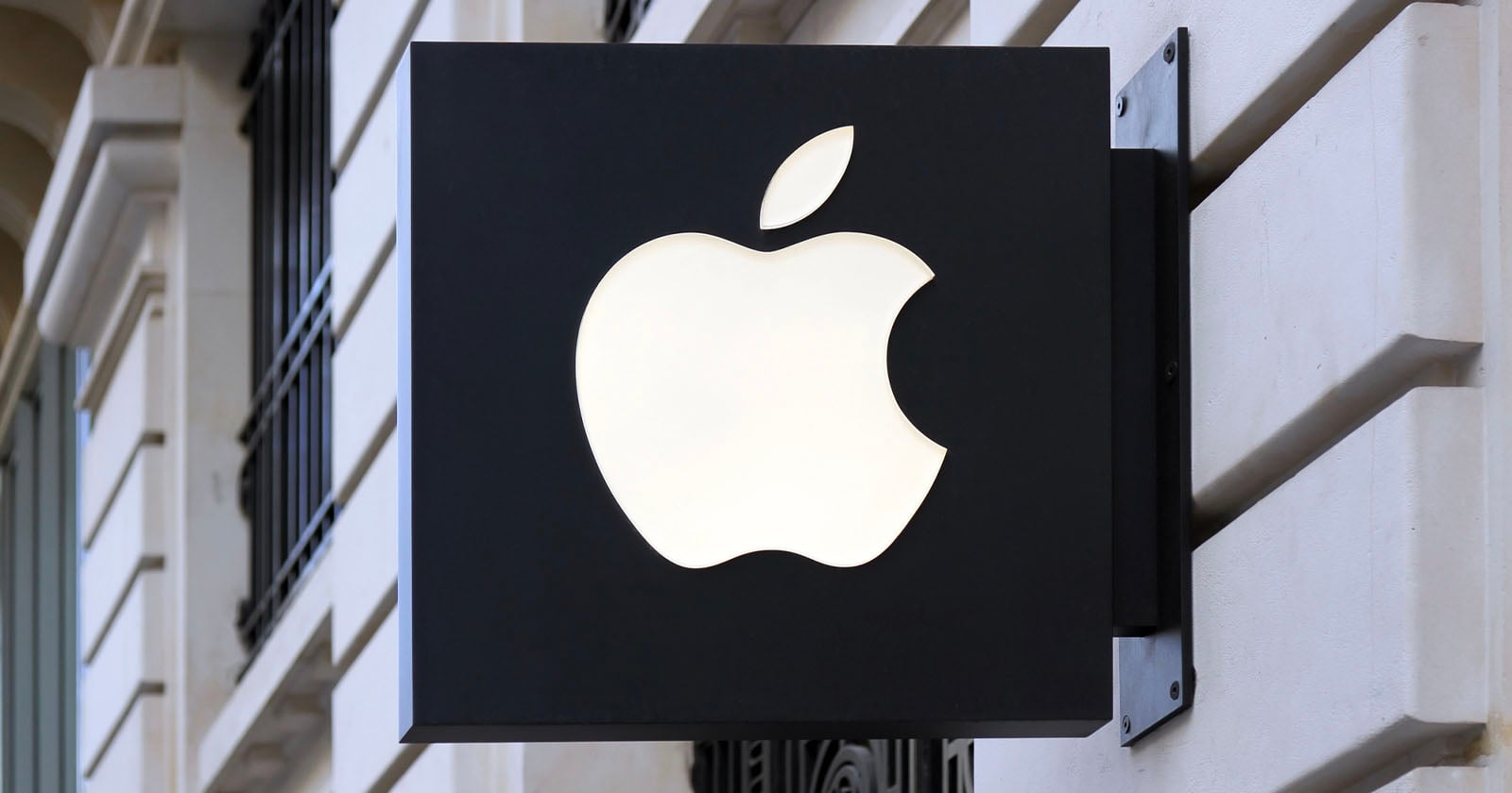 A black rectangular sign with a white, glowing Apple logo is mounted on the exterior wall of a building. The sign is attached with metal brackets and the building has light-colored stone and decorative window bars.