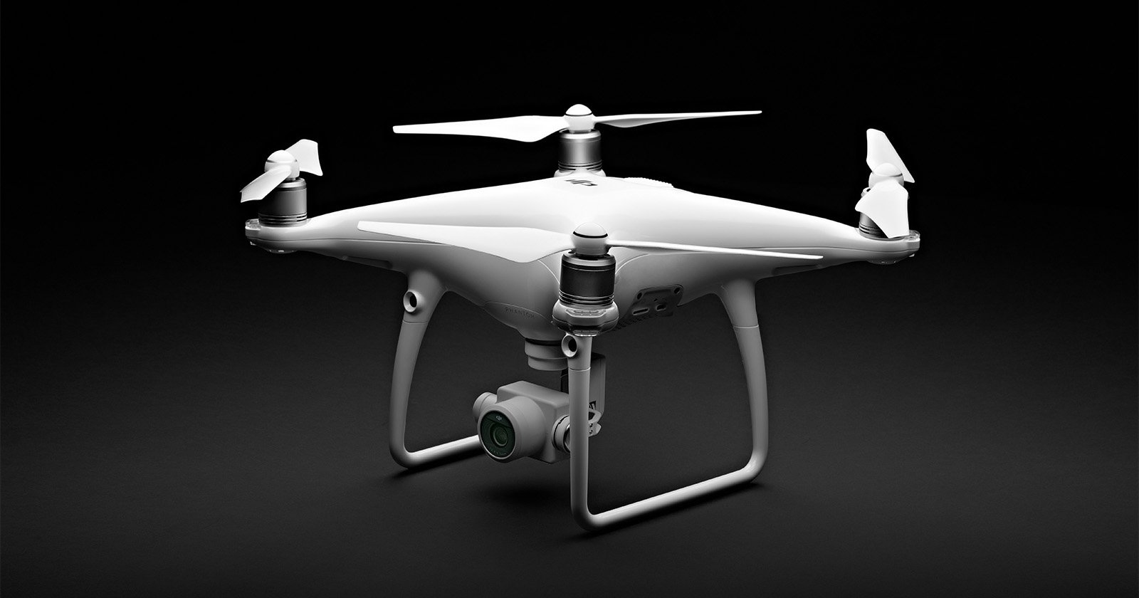 A white quadcopter drone with four propellers and a camera mounted underneath, shown against a dark background. The drone has a sleek, modern design with sturdy landing legs.